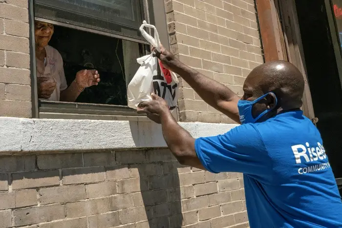 A worker from Citymeals on Wheels—he is wearing a mask—gives a bag of food to a woman through an open window from the sidewalk